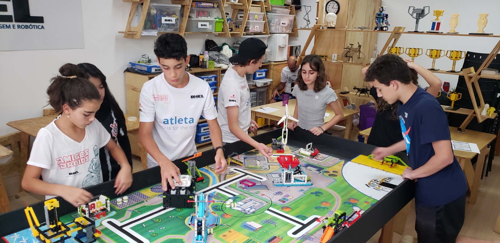Equipe de estudantes da DHEL ao redor da mesa de missões da FIRST LEGO League, com robôs LEGO e troféus ao fundo